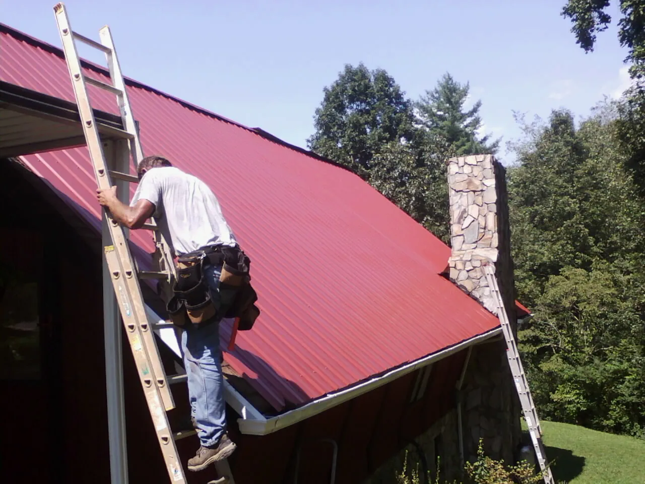 Roofer on a ladder beside a red metal roof and stone chimney — Soffit & Fascia Repair by Roofing Brothers in Condon